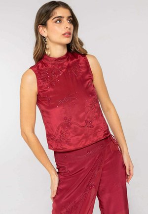 Mujer con top de satén rojo sin mangas y falda a juego con bordado floral de cuentas, mirando hacia un lado con el pelo largo y ondulado y pendientes de aro.