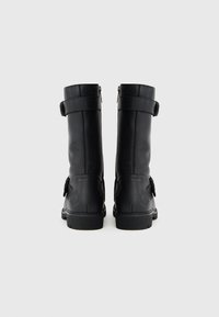 Um par de botas de couro pretas até à meia panturrilha com fivelas e fechos de correr, vistas pela parte de trás sobre um fundo branco liso.