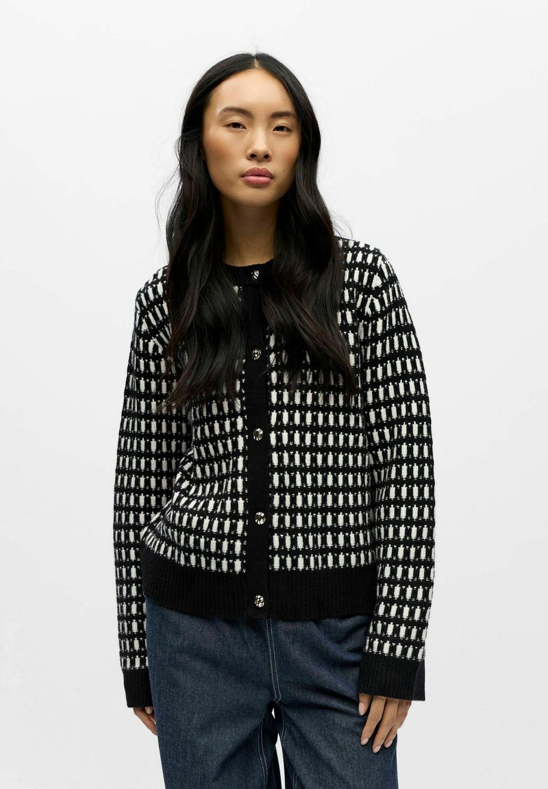 Melnā un baltā rakstu džemperis ar ribotu apakšmalu un piedurknēm, ar spīdīgām pogām priekšpusē. Strukturēta, adīta auduma.