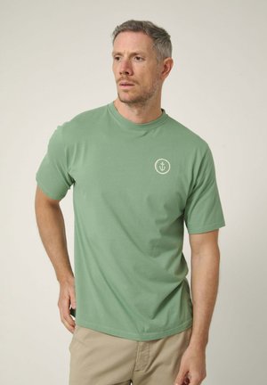 Hombre con camiseta verde claro con un pequeño logo blanco de ancla en el pecho izquierdo, pantalones beige, de pie frente a un fondo liso.