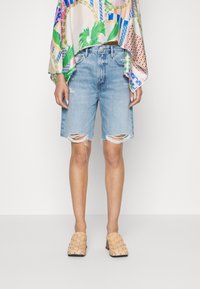 Pantalones cortos de mezclilla azul claro con dobladillos deshilachados y desgaste, combinados con una camiseta oversized de estampado colorido y zapatos de lona beige tipo slip-on.