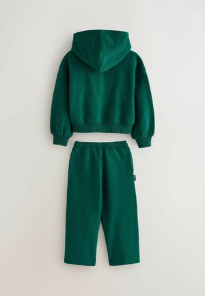 Zeleni bombažni pulover s kapuco in rebrastimi manšetami, v kombinaciji z usklajenimi zoženimi trenirkami. Obe stvari imata mehak teksturo in sproščen kroj.