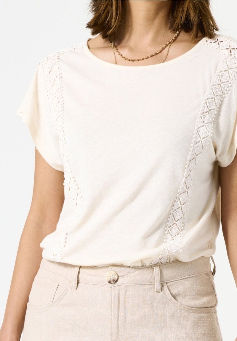 Žena v krémovém topu s krátkým rukávem a krajkovými detaily na ramenou a po stranách, vmáčknutém do béžových kalhot s vysokým pasem a knoflíkovým zapínáním.