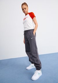 Sive Nike trenirke s vezicom, uparen s bijelom i crvenom majicom s raglan rukavima koja prikazuje grafiku. Bijele tenisice upotpunjuju odjevnu kombinaciju.