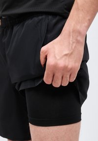 Κοντινό πλάνο ενός χεριού που ξεκουράζεται στην τσέπη μαύρων αθλητικών σορτς πολλαπλών στρώσεων που φοριούνται πάνω από κοντά σορτς συμπίεσης.