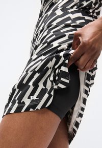 Αθλητική φούστα σε ασπρόμαυρο με ζιγκ ζαγκ σχέδιο, κατασκευασμένη από απαλό ύφασμα. Διαθέτει στενά μαύρα σορτς από κάτω και διακριτικό λογότυπο.