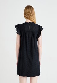 Μαύρο φόρεμα με κοντά λυμένα μανίκια, με κηλίδες σε όλη την ε�πιφάνεια και κουμπί στο πίσω μέρος. Χαλαρή εφαρμογή με πτυχές στη μέση.