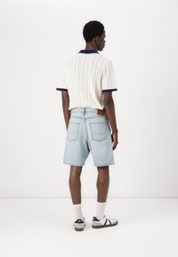 Polo perforado de manga corta, blanco con ribete azul marino, combinado con pantalones cortos de mezclilla azul claro que cuentan con dos bolsillos traseros y un parche de cuero marrón.