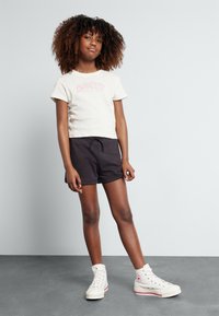 Lys beige avskåret t-skjorte med "ROXY Siden 90" i rosa, sammen med svarte shorts og hvite høye sneakers med røde detaljer.