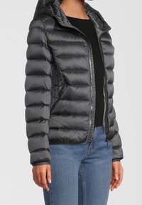 Svart puffjacka med huva, med dragkedjestängning, quiltat mönster och ribbade mudd, i kombination med en svart topp och blå jeans.