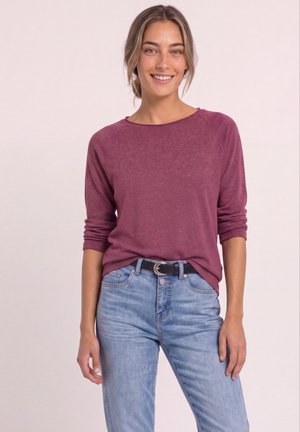 Ung kvinde smilende, iført en bordeaux langærmet top stoppet ned i lyseblå højttaljede jeans med sort bælte.