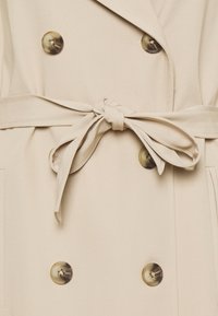 Beige trenchcoat met een strikceintuur, dubbelbreasted ontwerp, grote donkere knopen en een gladde textuur. Voorzien van opgestikte zakken aan de voorkant.