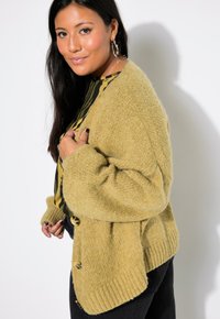 Hořčicově žlutý pletený svetr s širokými rukávy, oversized střihem, žebrovanými manžetami a dřevěnými knoflíky. Spárován s proužkovaným trikem.