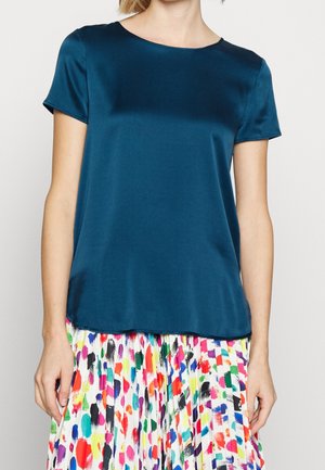 Kvinde iført en kortærmet, mørk grønblå satinbluse parret med en hvid plisseret nederdel med flerfarvede malingslignende mønstre.