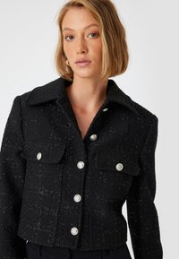 Veste noire courte en tissu texturé, dotée d'un col, de fermetures à bouton argenté et de poches à rabat à l'avant.