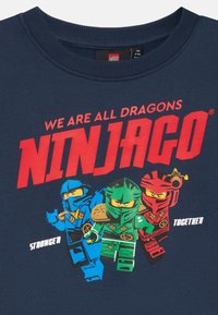 Tmavě modré tričko z látky s postavičkami LEGO Ninjago v modré, zelené a červené barvě. Velký červený nápis zní "VŠICHNI JSME DRACI NINJAGO."