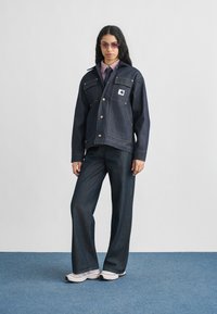 Σκούρο τζιν μπουφάν με μυτερό γιακά, εμπρόσθιες τσέπες και χρυσά κουμπιά, συνδυασμένο με τζιν παντελόνι φαρδιάς γραμμής που ταιριάζει και λευκά αθλητικά παπούτσια.