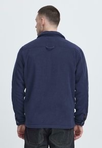 Chaqueta de forro polar azul marino con un alto cuello, puños abotonados y un lazo en el cuello. Presenta una textura suave y un ajuste entallado.