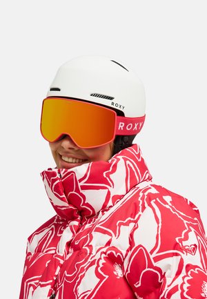 Lächende Person mit weißem Roxy-Skihelm, orange getönten Skibrillen und einer rot-weiß floral gemusterten Winterjacke.