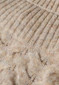 Tejido de punto beige con patrones de canalé y trenzas, que presenta una textura suave y peluda, en tonos variados de marrón y crema.