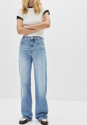 White short-sleeve shirt with black trim, paired with light blue high-waisted wide-leg jeans. The outfit is completed with dark shoes.