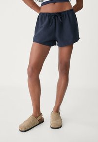 Námořnické modré bavlněné šortky s elastickým pasem a tkaničkou; doplněné béžovými semišovými dřeváky s kulatými hroty na podešvi.