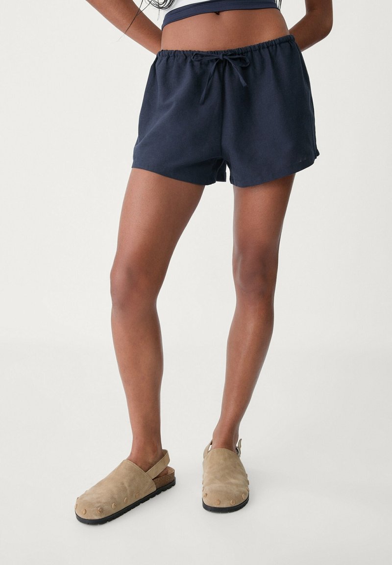 Námořnické modré bavlněné šortky s elastickým pasem a tkaničkou; doplněné béžovými semišovými dřeváky s kulatými hroty na podešvi.