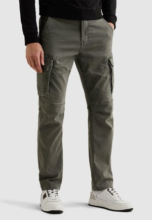 Muž oblečený v olivovo zelených nohaviciach typu cargo s viacerými vreckami, čierny top a biele tenisky, stojaci s rukami vreckách.