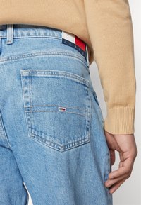 Vaaleansiniset farkut, joissa takatasku ja sen yläpuolella monivärinen logopatch. Hihassa näkyy beige neulepaita.