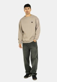 Beige Sweatshirt mit rundem Halsausschnitt und schwarzem Logo, kombiniert mit locker sitzenden dunkelgrünen Hosen und schwarzen Sneakers.