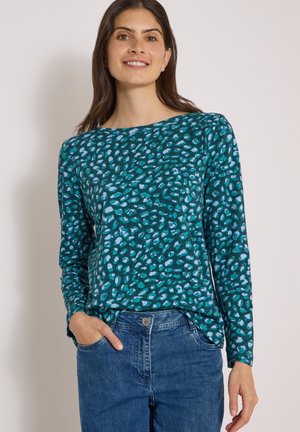 Femme portant un haut à manches longues à motifs bleu sarcelle et bleu avec une main dans la poche d'un jean bleu taille haute, souriant légèrement.