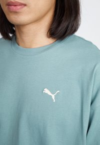 Tricou din bumbac teal, cu guler rotund, croială lejeră și un logo Puma brodat alb pe partea stângă a pieptului. Material moale, texturat.