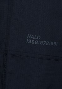 Granatowa tkanina o teksturze siatki. Zawiera wytłoczone napisy "HALO 196819721981." Szczegóły wyszywane wzdłuż szwów. Trwały materiał.