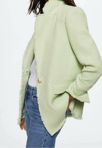 Jasnozielona, teksturowana marynarka z postrzępionymi krawędziami, zapinana na pojedynczy perłowy guzik, z bocznymi kieszeniami, zestawiona z białą koszulą i dżinsami.