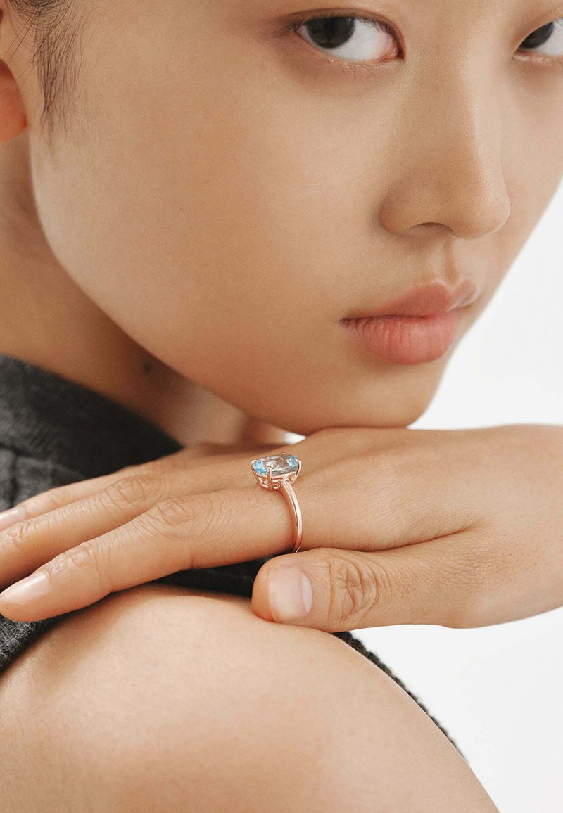 Anillo de oro rosado con una gran piedra preciosa azul clara, montada en una banda elegante y minimalista, exhibido en una mano contra un fondo neutro.