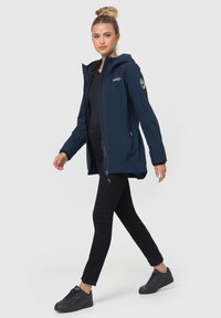 Chubasquero impermeable azul marino con capucha, cierre de cremallera y bolsillos, con un parche de logo en la manga izquierda. Combinado con pantalones negros y zapatillas.