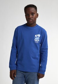 Blaues Langarm-T-Shirt mit weißem Miami-Themen-Graphic auf der linken Brust. Tragen mit blauen Jeans, Rundhalsausschnitt und genähten Bündchen.