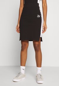 Donna che indossa un vestito Puma nero fino al ginocchio con pannelli laterali bianchi, calze bianche Puma e sneakers grigio chiaro Puma, in piedi su uno sfondo neutro.