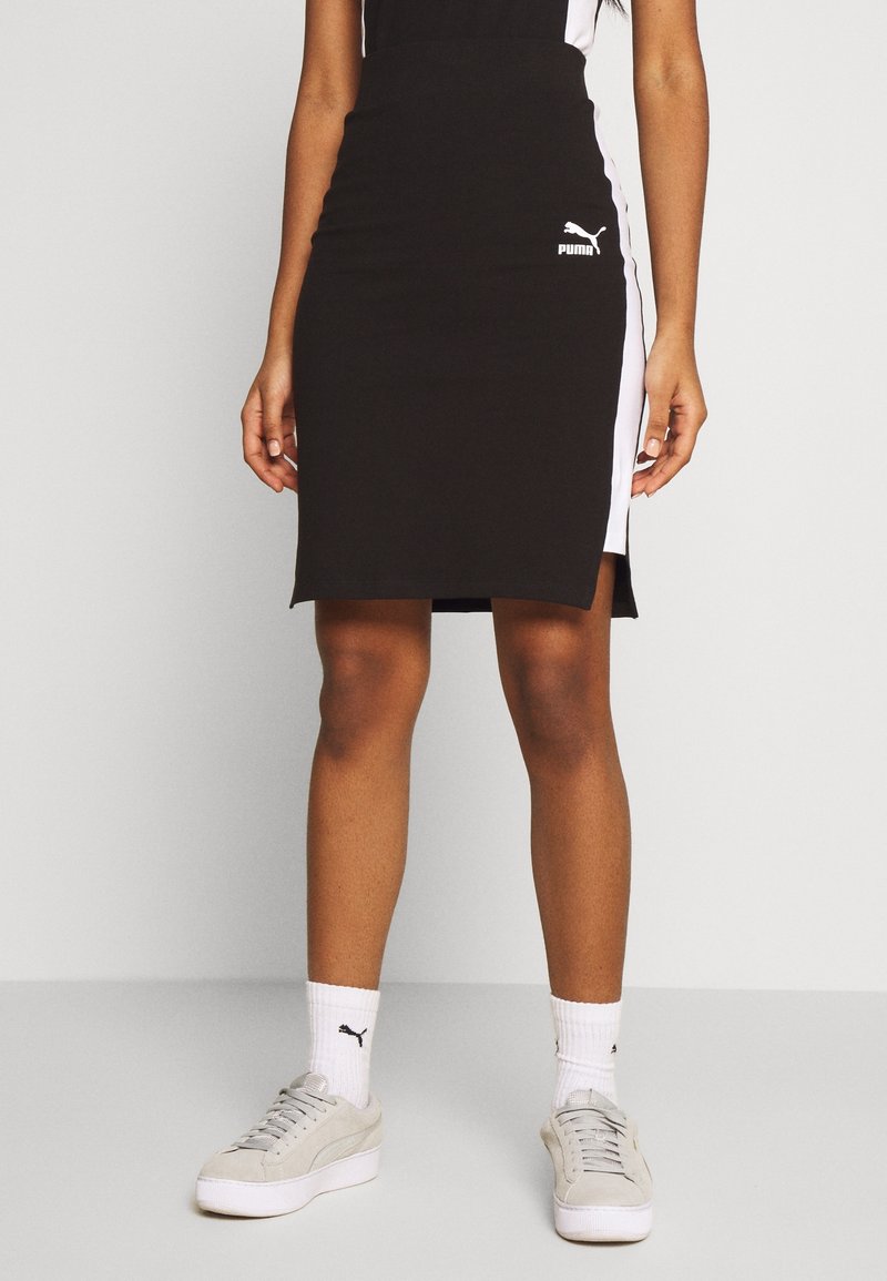 Donna che indossa un vestito Puma nero fino al ginocchio con pannelli laterali bianchi, calze bianche Puma e sneakers grigio chiaro Puma, in piedi su uno sfondo neutro.