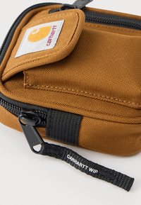 Καφέ υφασμάτινη θήκη με μαύρο φερμουάρ, μαύρη ετικέτα τραβήγματος φερμουάρ με την ένδειξη "Carhartt WIP" και μικρή μπροστινή θήκη με καπάκι και λογότυπο Carhartt.