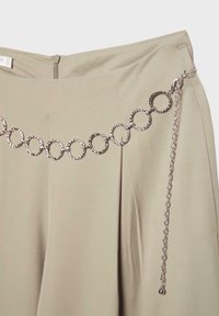 Pantalon beige clair avec une ceinture chaîne argentée composée de maillons circulaires martelés et une fermeture à chaîne ajustable sur le côté droit.