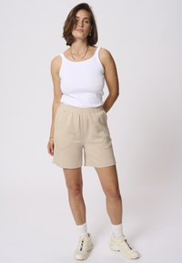 Shorts en beige avec taille élastique associés à un débardeur blanc. Le mannequin porte des chaussettes blanches et des baskets claires. Fond neutre.