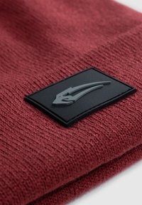 Beanie rosso a maglia caratterizzato da un tessuto testurizzato e un evidente patch in gomma nera con un design del logo stilizzato.