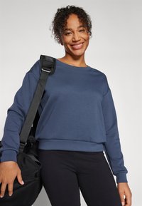 Felpa blu scuro con scollo ampio, maniche lunghe e vestibilità rilassata. Abbinata a leggings neri e a una borsa da viaggio nera.