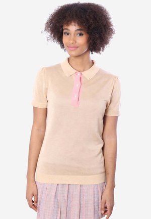 Giovane donna con capelli ricci che indossa una polo beige a maniche corte con patta rosa e una gonna plissettata pastello, sorridente dolcemente.