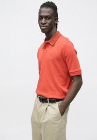 Polokošile s krátkým rukávem v živé oranžové barvě, texturovaná látka, s límečkem a malým vyšívaným detailem na hrudi; ve spojení s béžovými kalhotami.