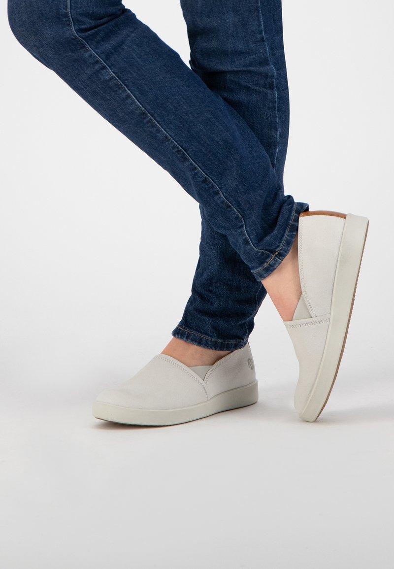 Chaussures à enfiler gris clair en tissu lisse, avec un bout arrondi et des accents élastiques sur les côtés, associées à un jean en denim bleu foncé.
