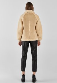 Kobieta w beżowej polarowej kurtce z widocznym szyciem, czarnych skórzanych, krótkich spodniach i czarnych botkach za kostkę, stojąca tyłem.