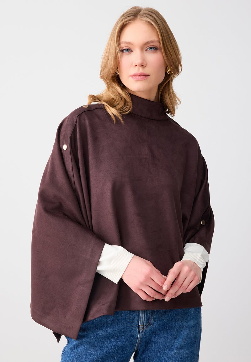 Femme blonde portant un poncho marron foncé à boutons dorés par-dessus un haut blanc à manches longues et un jean bleu, debout devant un fond uni.
