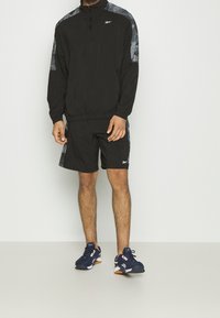 Chándal atlético negro con chaqueta de medio cierre, mangas cortas y pantalones cortos. Acentos grises en las mangas. Combinado con zapatillas navy.